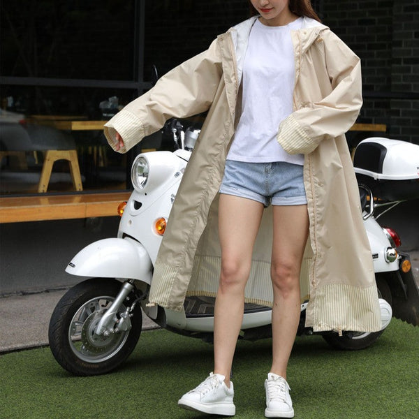 Une femme pose en extérieur devant un scooter blanc. Elle porte un tee-shirt blanc, un short en jean et des baskets blanches. Elle porte également une veste imperméable longue coupe vent beige à rayures blanches sur le bas et les poignets.