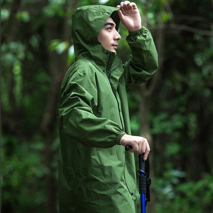 Un homme pose dans la forêt avec une veste longue imperméable kaki avec capuche et dos extensible. Il porte un batôn de marche bleu dans une main.