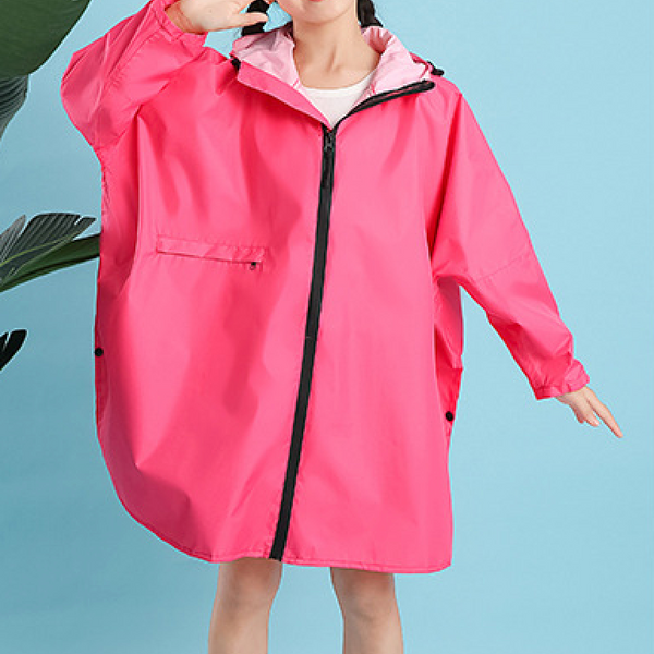 Une enfant pose devant un fond bleu avec une plante à côté d'elle. Elle porte un tee-shirt blanc et une veste imperméable rose