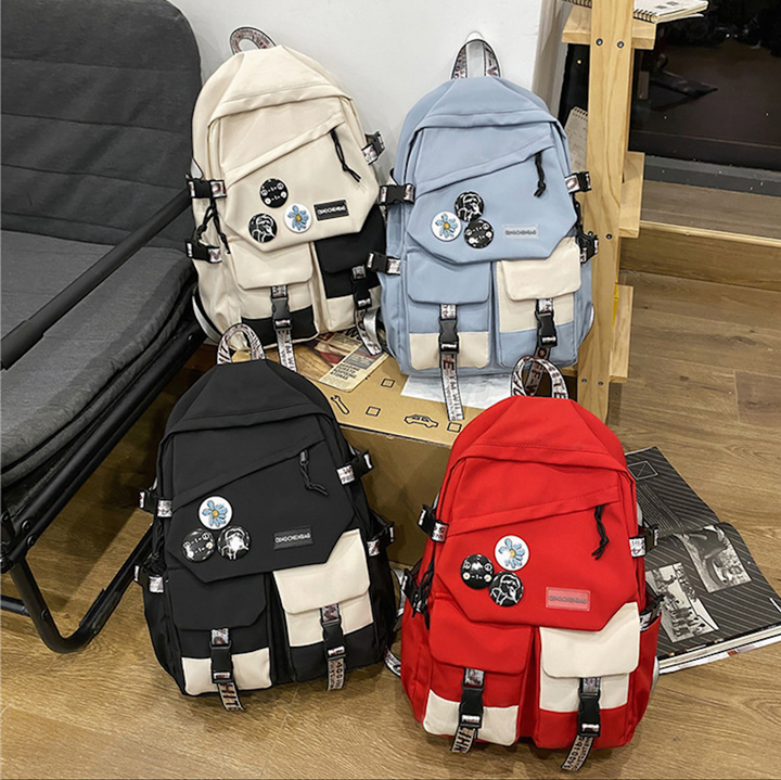 Dans un salon avec parquet et canapé gris, 4 sacs est posés sur un carton. Les sacs à dos sont imperméables et étanches, multipoches et patchs. Il y en a blanc, bleu,rouge et noir. 