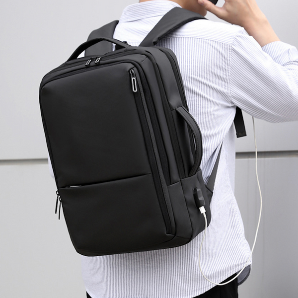 Un homme pose dans la rue avec une chemise blanche à fines rayures bleues et un pantalon noir. Il porte aussi un sac à dos simple noir. Il est étanche et a des ports USB.