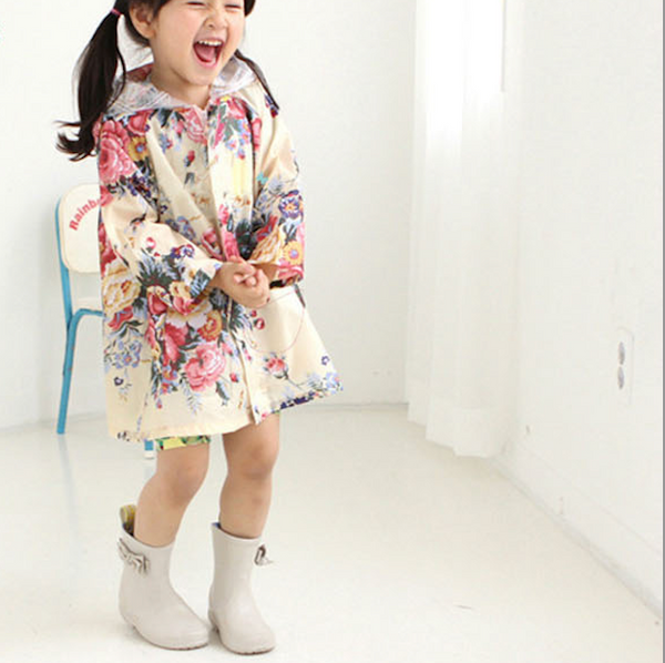 Une petite fille porte une veste imperméable large à capuche blanc avec un motif floral coloré. Elle porte aussi des bottes de pluie beige. 