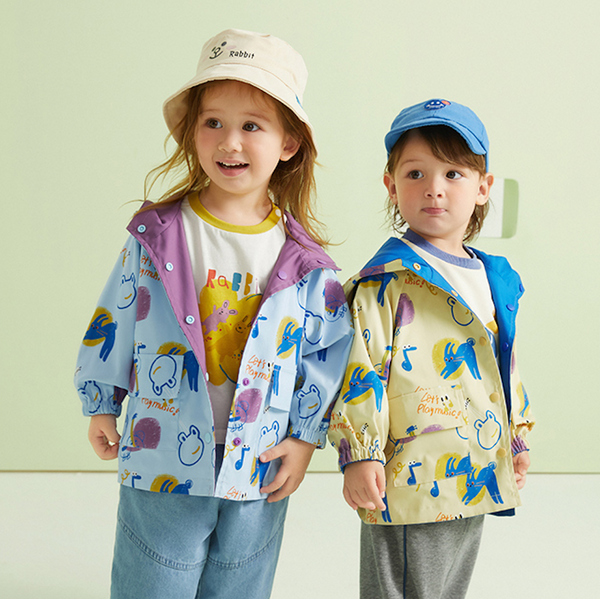 Deux enfants posent devant un fond vert. Ils portent tous les deux une vestes imperméables à capuche réversible. Il y a un côté uni et l'autre côté a un motif dessin animaux. 