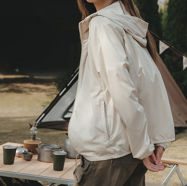 Une femme pose devant une table de camping et une tente. Elle porte un pantalon cargo kaki et une veste imperméable coupe-vent léger anti-UV beige. 