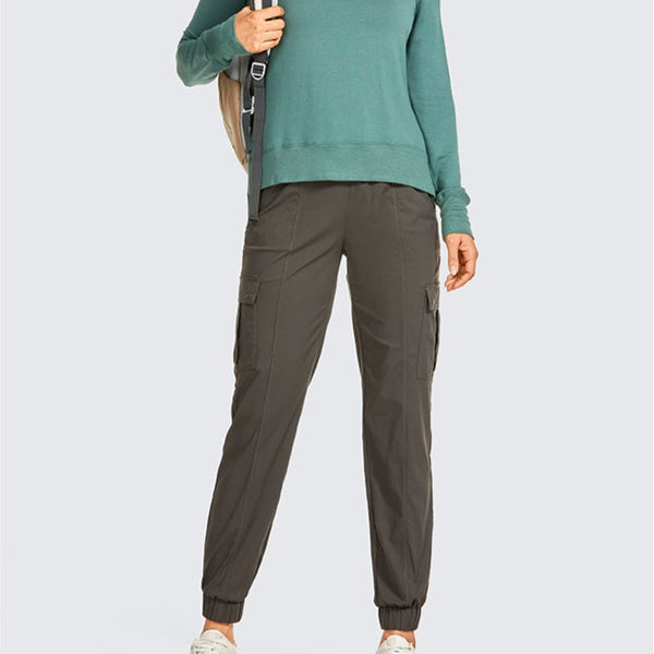 Une femme pose devant un fond gris. Elle porte un pull vert, un sac à dos et un pantalon imperméable jogging cargo kaki.