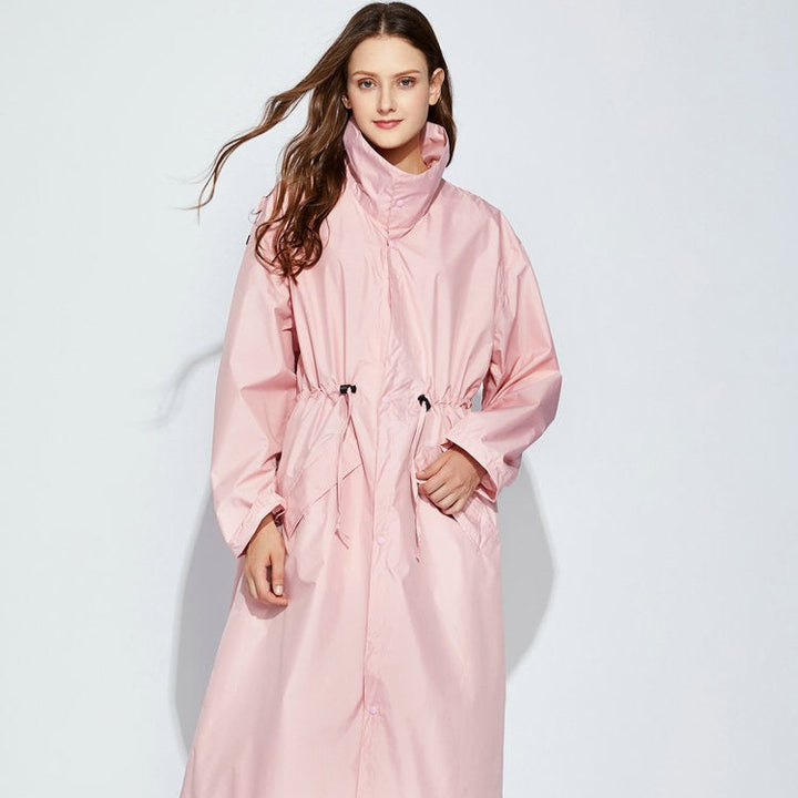 Une femme pose sur un fond gris clair. Elle est brune aux cheveux long et porte une longue veste imperméable longue rose cintrée avec col montant. 