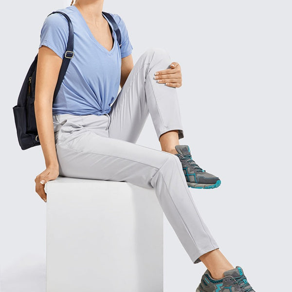 Une femme pose sur un cube. Elle porte un tee-shirt bleu, un sac à dos noir, des baskets de randonnée et un pantalon imperméable léger taille haute blanc. 