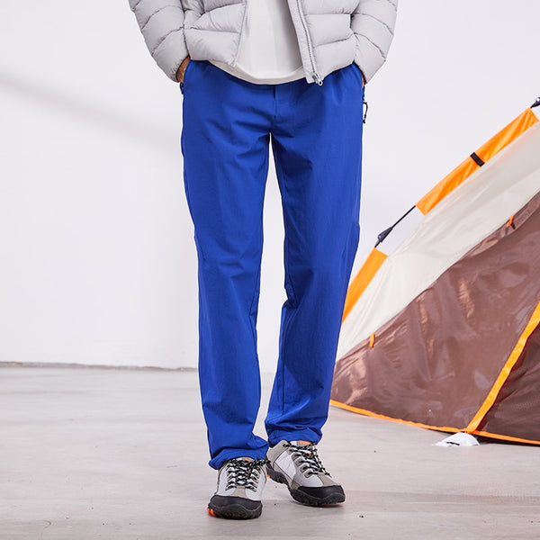 Un homme pose devant une tente. Il porte une doudoune grise, des chaussures basses de montagne et un pantalon imperméable de randonnée bleu. 