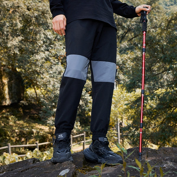 Un homme pose dans la nature. Il porte un tee-shirt noir, une paire de baskets noirs. Il porte un bâton de marche dans la main. Il porte aussi un pantalon imperméable de randonnée bicolore gris et noir. 