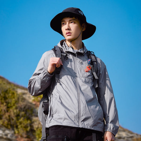 Un homme pose en pleine nature avec un bob noir, un pantalon noir , un sac à dos et une veste imperméable grise coupe vent légère et respirante. 