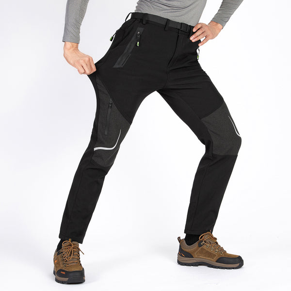Un homme pose devant un fond blanc. Il porte des chaussures de montagne marron, un tee-shirt manches longues gris, un pantalon noir imperméable avec bandes réfléchissantes sur les genoux.