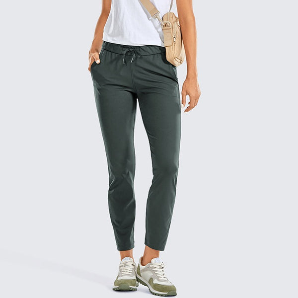 Une femme pose devanrt un fond gris. Elle porte un tee-shirt blanc, des baskets, un sac en bandoulière et un pantalon imperméable ajusté vert olive. 