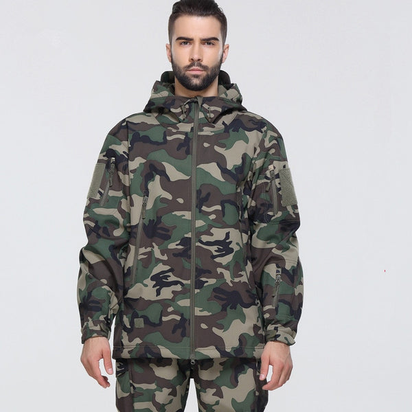 Un homme pose devant un fond gris. Il porte une veste imperméable camouflage kaki et un pantalon assorti.