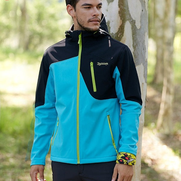 Un homme pose dans la nature. Il porte un pantalon bleu marine et une veste imperméable coupe-vent bicolore noir et bleu turquoise avec des fermetures vert fluo. 