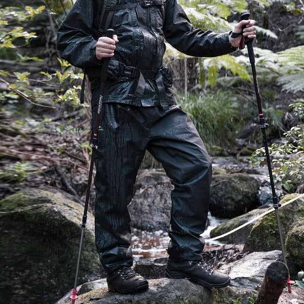 Un homme pose en pleine nature. Il porte une veste imperméable noir, un sac à dos. Il tient dans chaque main un bâton de marche et porte aussi un pantalon de pluie imperméable noir.