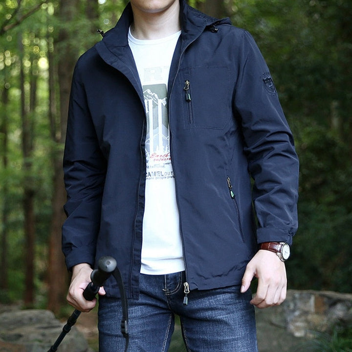 Un homme pose dans la forêt avec un tee-shirt blanc, un jean et une veste imperméable bleu foncé avec poches zipées. La veste est imperméable. Il porte aussi un bâton dans une des mains. 