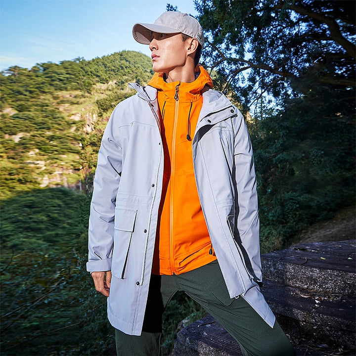 Un homme pose dans la nature. Il porte une casquette beige, une veste orange, un pantalon kakie et une veste grise mi-longue à capuche.