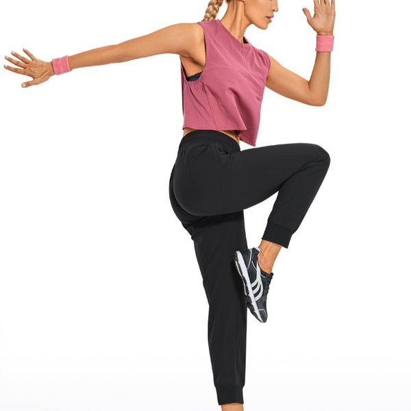 Une femme pose en faisant du sport. Elle porte un tee-shirt rose, des baskets noires et blanches et un pantalon imperméable et étanche de type jogging léger noir. 