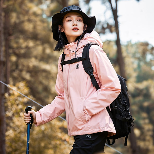 Une femme pose dans la forêt. Elle porte un bob noir, des bâtons, un sac à dos noir, un pantalon noir ainsi qu'une veste imperméable rose coupe-vent légère. 