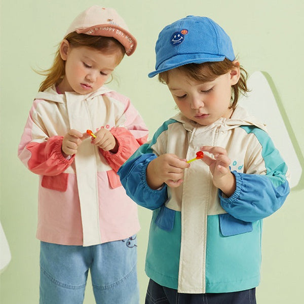 Deux enfants posent devant un fond vert. Les enfants portent des jeans, des casquettes et des vestes imperméables. Les vestes sont colorées. La petite fille en porte une beige et rose et le garçon une bleu et beige. 