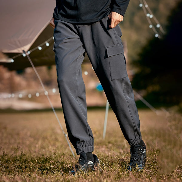 Un homme pose dans la nature. Il y a une tente et des guirlandes lumineuses derrière lui. Il porte un haut à manches longues noir, des baskets noires et un pantalon cargo gris anthracite imperméable doublé polaire. 