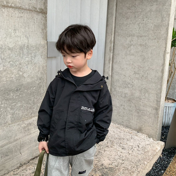 Un petit garçon pose dans la rue. Il porte un jogging gris, un sac kaki et un tee-shirt noir. Il porte aussi une veste noir imperméable coupe-vent à capuche.
