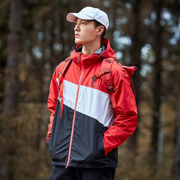 Un homme pose en pleine forêt. Il porte une casquette blanche, un pantalon noir, un sac à dos rouge et une veste imperméable coupe-vent tricolore rouge, bleu et blanche.