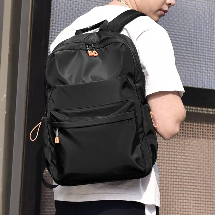 Une homme pose avec un sac à dos noir à poche zippé sur le devant. Le sac à dos est imperméable et muni de port USB. 