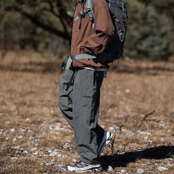 Un homme pose dans la nature. Il porte une veste marron, un sac à dos, des chaussures de randonnée et un pantalon imperméable de randonnée large gris. 