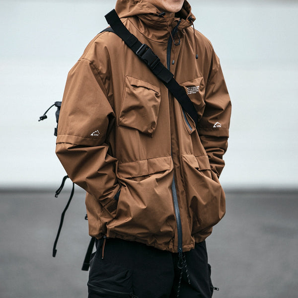 Un homme pose dans la rue. Il porte un sac en bandoulière, un pantalon noir et une veste imperméable multipoches marron. 