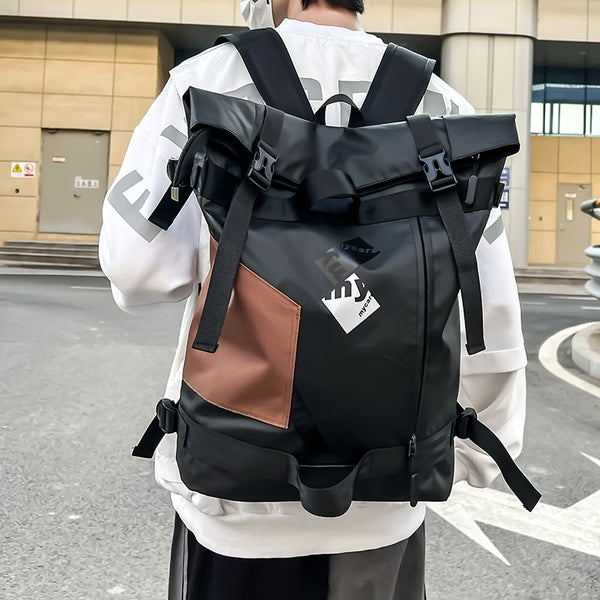 Un homme pose dans la rue avec un haut manches longues blanc et un baggy noir et gris. Il porte un grand sac à dos noir imperméable avec rabat et poche patchwork camel. 