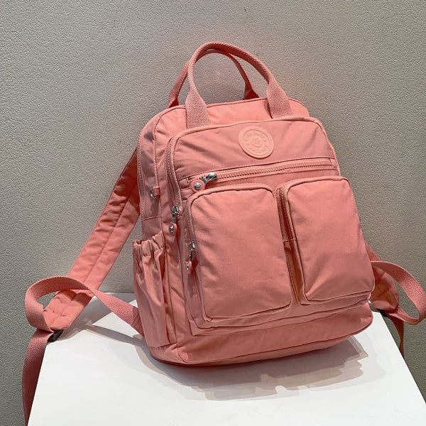 Un sac à dos est posé sur une petite table blanche devant un mur blanc. Le sac à dos est rose avec de nombreuses poches sur le devant. Il a une poignée sur le dessus et des bretelles. 