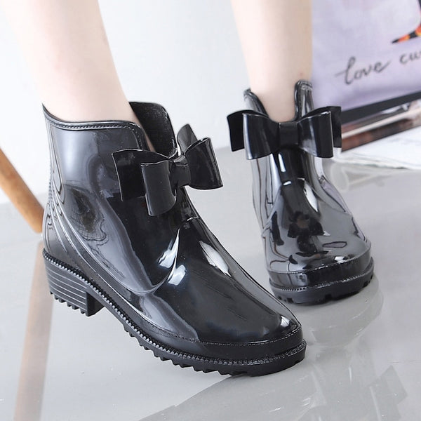Une femme porte des bottes de pluie basses avec un noeud sur le devant. On voit uniquement ses pieds. Les bottes sont noires. 