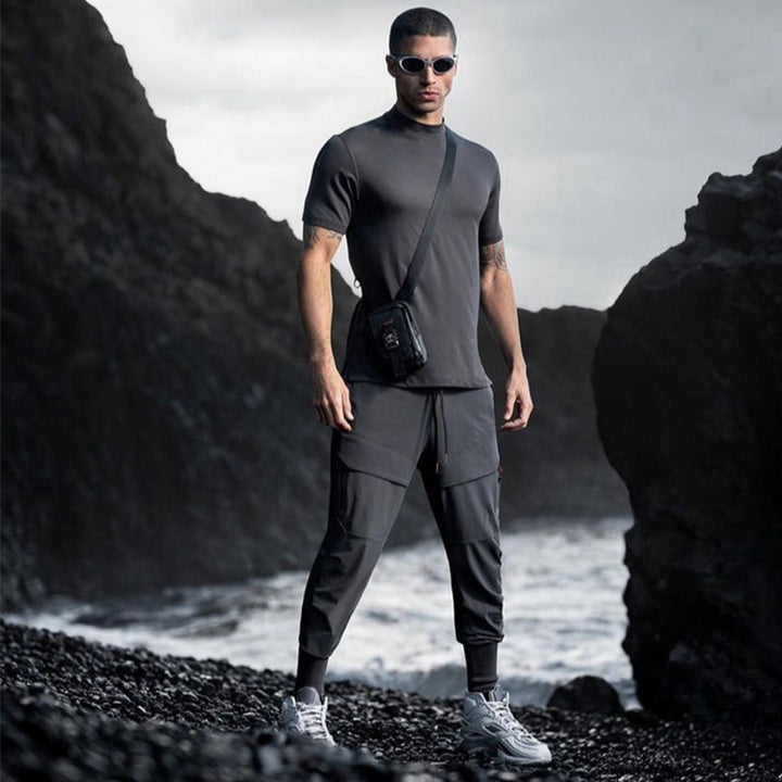 Un homme pose sur une plage. Il porte un tee-shirt gris, des baskets et un pantalon imperméable jogging gris. Il porte des lunettes de soleil et une sacoche en bandoulière.