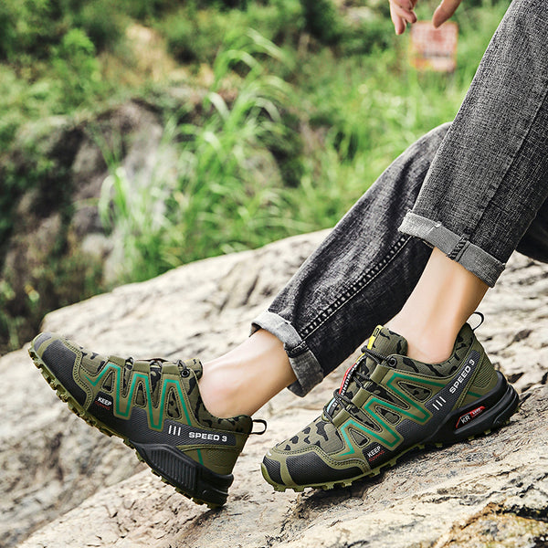 Un homme pose sur un rocher en pleine nature ; On ne voit que ses jambes et ses pieds. Il porte un jean gris retroussé et une paire de chaussures de randonnée imperméables vertes à motif camouflage.