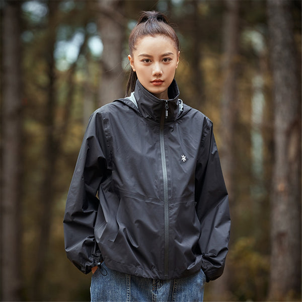 Une femme pose en forêt dans un pantalon en jean et avec une veste courte imperméable coupe-vent large noir. Elle a les cheveux bruns et une queue de cheval.