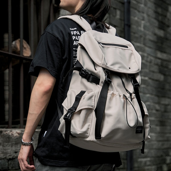 Un homme pose dans la rue avec un sac à dos imperméable étanche avec rabat et cordon de serrage. Le sac est beige avec et l'homme est habillé en noir. 