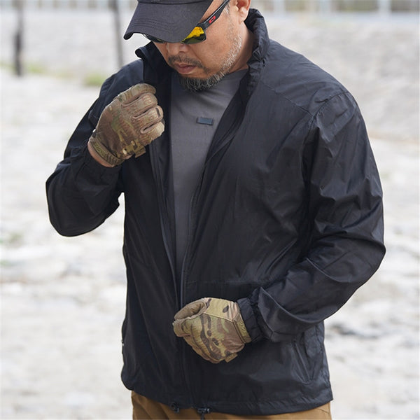 Un homme pose à l'extérieur avec une veste imperméable légère coupe vent noire. Il porte des gants motif camouflage, un pantalon camel, un tee-shirt gris anthracite et une casquette noire. Il a aussi des lunettes de soleil.