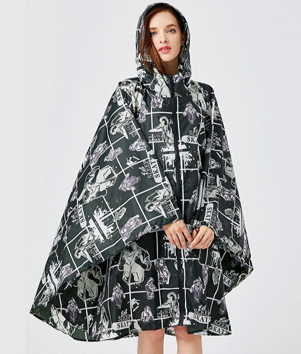 Une femme pose devant un fond gris dans une veste imperméable poncho noir avec motif blanc. Elle est brune aux yeux bleus