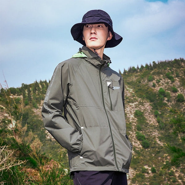 Un homme pose dans la nature. Il porte un bob noir, un pantalon noir, une veste imperméable kaki avec une capuche vert fluo. 