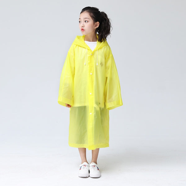 Une enfant pose devant un fond gris. Elle est brune aux cheveux attachés. Elle porte un tee-shirt blanc, des baskets blanches ainsi qu'une veste imperméable longues à manches larges jaune. 