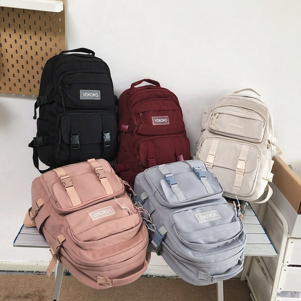 5 sacs sont posés sur une table. C'est le même modèle un sac à dos imperméable avec multipoches à clip. Seul la couleur diffère. Il y en a un noir, un rouge, un beige, un rose et un bleu gris.