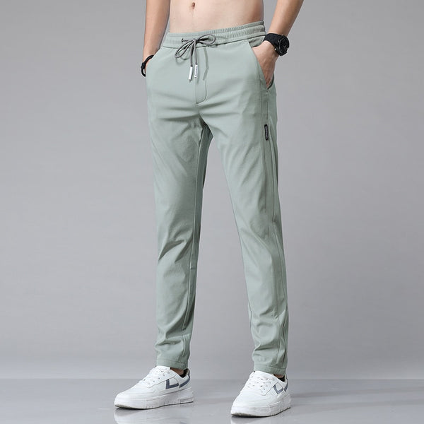 Un homme pose devant un fond gris. Il porte des baskets blanches et un pantalon imperméable léger et respirant vert clair. 