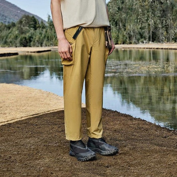 Un homme pose dans la nature. Il porte un tee-shirt jaune clair, un pantalon beige imperméable coupe droite large et des chaussures noires