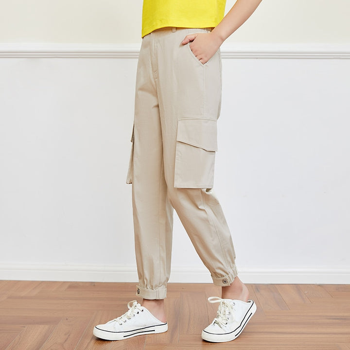 Une femme pose dans une pièce aux murs blancs avec parquet au sol. Elle porte un tee-shirt jaune, des baskets blanches et un pantalon imperméable cargo beige.