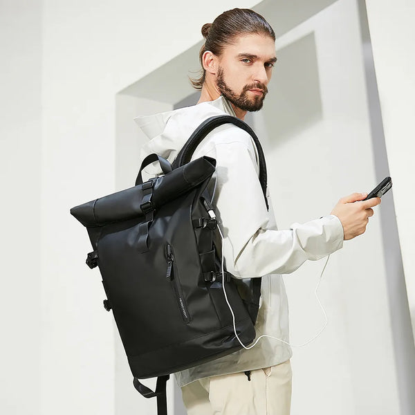 Un homme pose avec un sac sur le dos. Le sac à dos est noir avec rabat et port USB. Il est imperméable.