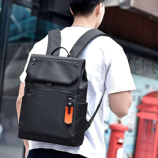 Un homme pose dans la rue avec un tee-shirt blanc et un short noir. Il a un sac à dos étanche et imperméable noir. 