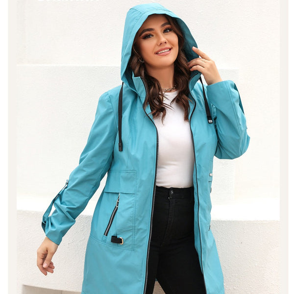 Une femme pose à l'extérieur devant des murs blanc. Elle est brune avec des cheveux ondulés. Elle porte un tee-shirt blanc, un jean taille haute noir et une veste mi-longue imperméable bleu. 