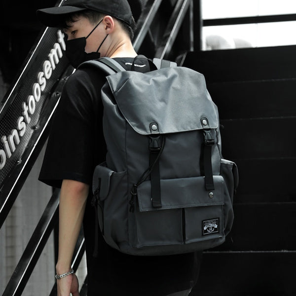 Un homme pose dans une cage d'escalier. Il porte une casquette noire, un masque noir, un tee-shirt noir et un sac à dos imperméable et étanche gris à rabat.