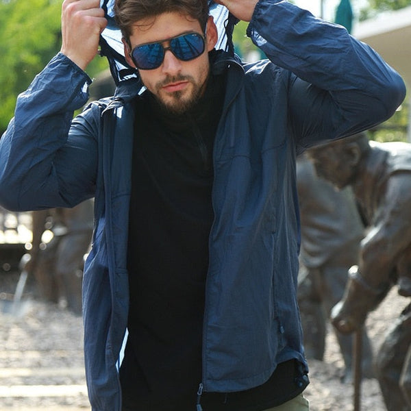 Un homme pose dans la rue devant des statues. Il porte des lunettes de soleil, un pull noir, un pantalon kaki et une veste imperméable anti UV bleu ultra fine et légère.